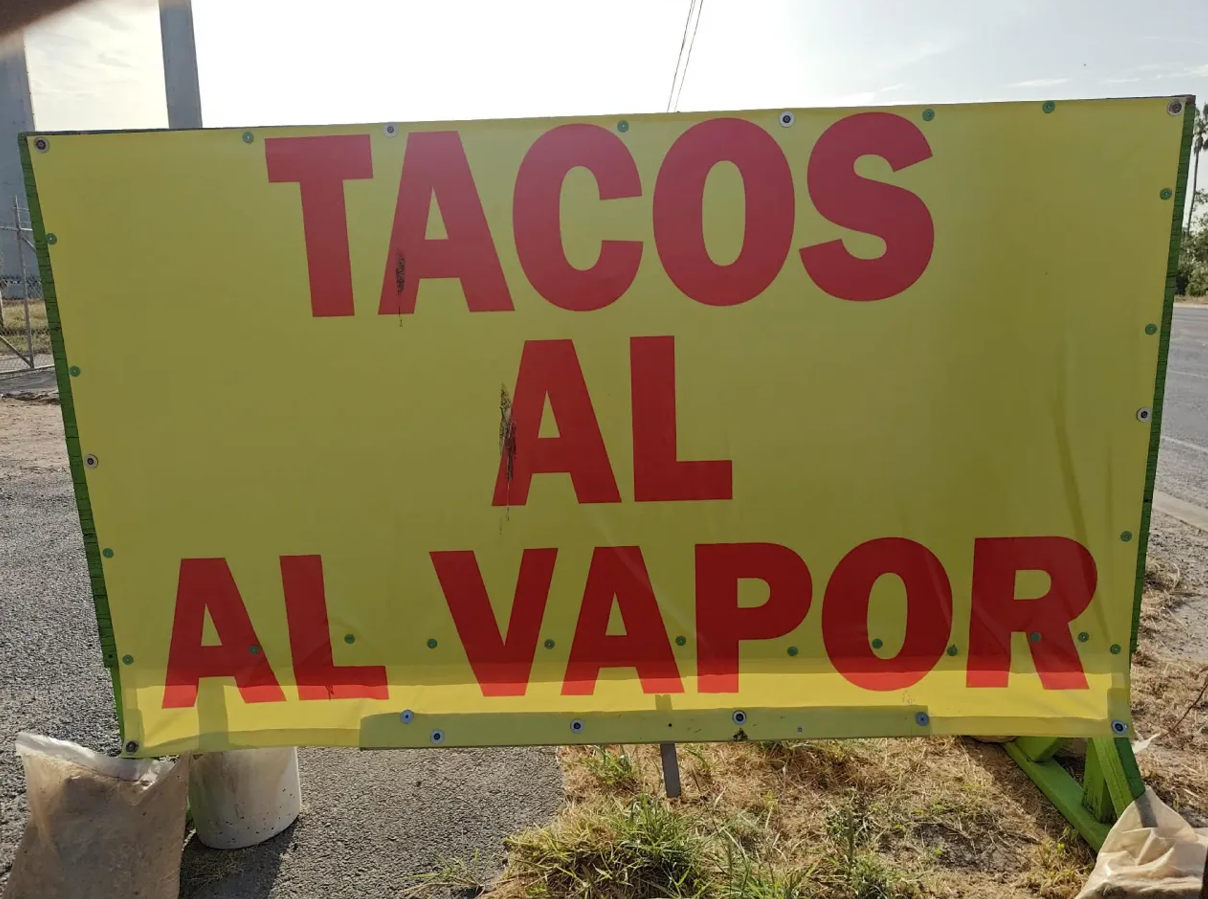 Lona amarilla de Tacos al Vapor sostenida por un marco verde a la orilla de la carretera.